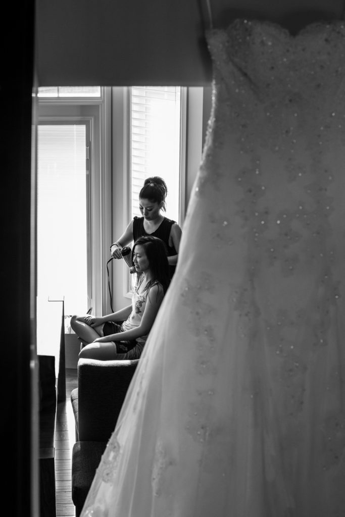 Bride sitting and getting her hair done with her wedding dress hanging in the foreground
